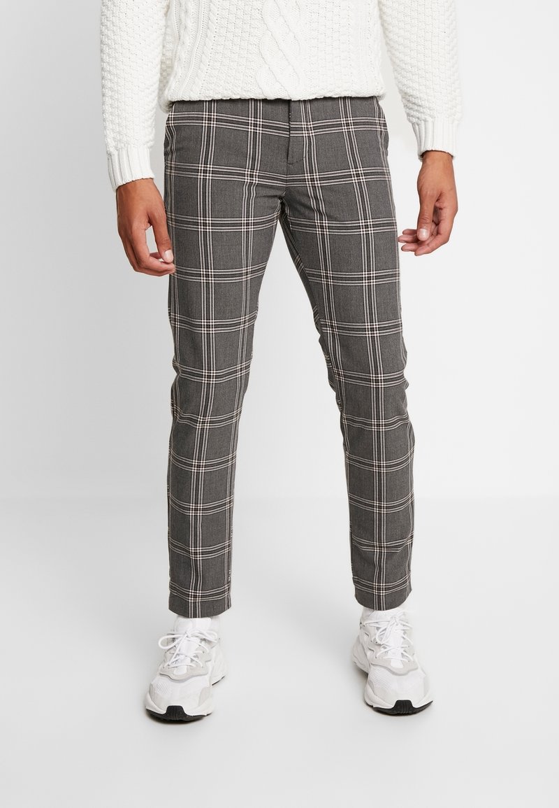 Homme portant un pantalon slim à carreaux gris, un pull en maille blanc et des baskets blanches, debout devant un fond uni.