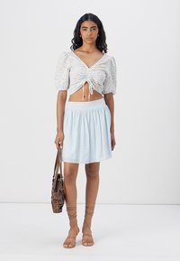 Hvitt blomster crop top med pufferm, kombinert med et lyseblått skjørt. Modellen holder en brun veske og har på seg beige sandaler med stropper.