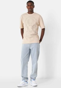 Camiseta gráfica beige de manga corta, con un diseño circular de sol y texto. Combinada con jeans de mezclilla azul claro y zapatillas blancas.
