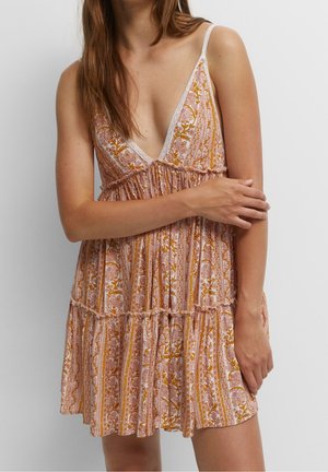 Robe d'été sans manches à col en V avec des rayures florales roses, blanches et moutarde, portée par une femme aux longs cheveux bruns sur un fond uni.