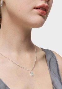 Collar de cadena de plata con un colgante en forma de oso sobre una superficie suave, llevado sobre una blusa gris de textura suave y escote drapeado.