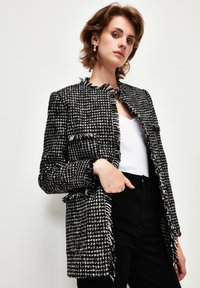 Černobílý tvídový blazer s třásněmi, límcem s výstřihem a předními kapsami, nošený přes bílý top a tmavé kalhoty.