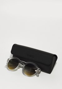 Ochelari de soare rotunzi cu cadru transparent și lentile închise la culoare cu gradient, așezați pe un toc texturat negru, embosat cu logo-ul "GUESS".