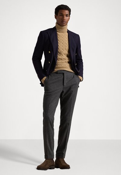 Donkerblauwe double-breasted blazer, tan kabelgebreide coltrui, grijze geplooide broek en bruine suède enkellaarzen.