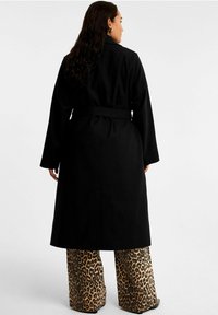 Zwarte lange jas met een ceintuur in de taille, voorzien van brede mouwen en een rechte snit, gecombineerd met een leopardprint wijde broek.
