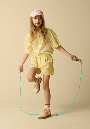 Junges Mädchen in gelbem Outfit und pinker Mütze springt Seil mit einem erhobenen Fuß vor einem einfarbigen beigen Hintergrund.