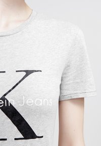 Osoba v světlém šedém tričku s velkým černým "K" a částečným nápisem "lein Jeans" viditelným z blízka.