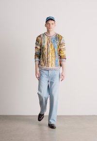 Monivärinen neule, jossa on aaltoilevia kuvioita ja pyöreä kaula-aukko, yhdistettynä vaaleansinisiin leveälahkeisiin farkkuihin ja tummiin kenkiin. Päässä on lippalakki.