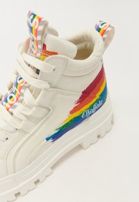 Zapatilla blanca alta de cuero con un diseño de pinceladas de arcoíris coloridas en el costado y un logo en azul. Suela blanca gruesa.