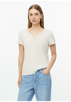 Femme aux cheveux lisses châtain clair portant un t-shirt crème à manches courtes avec col en V et un jean bleu clair, une main dans la poche, sur fond blanc.