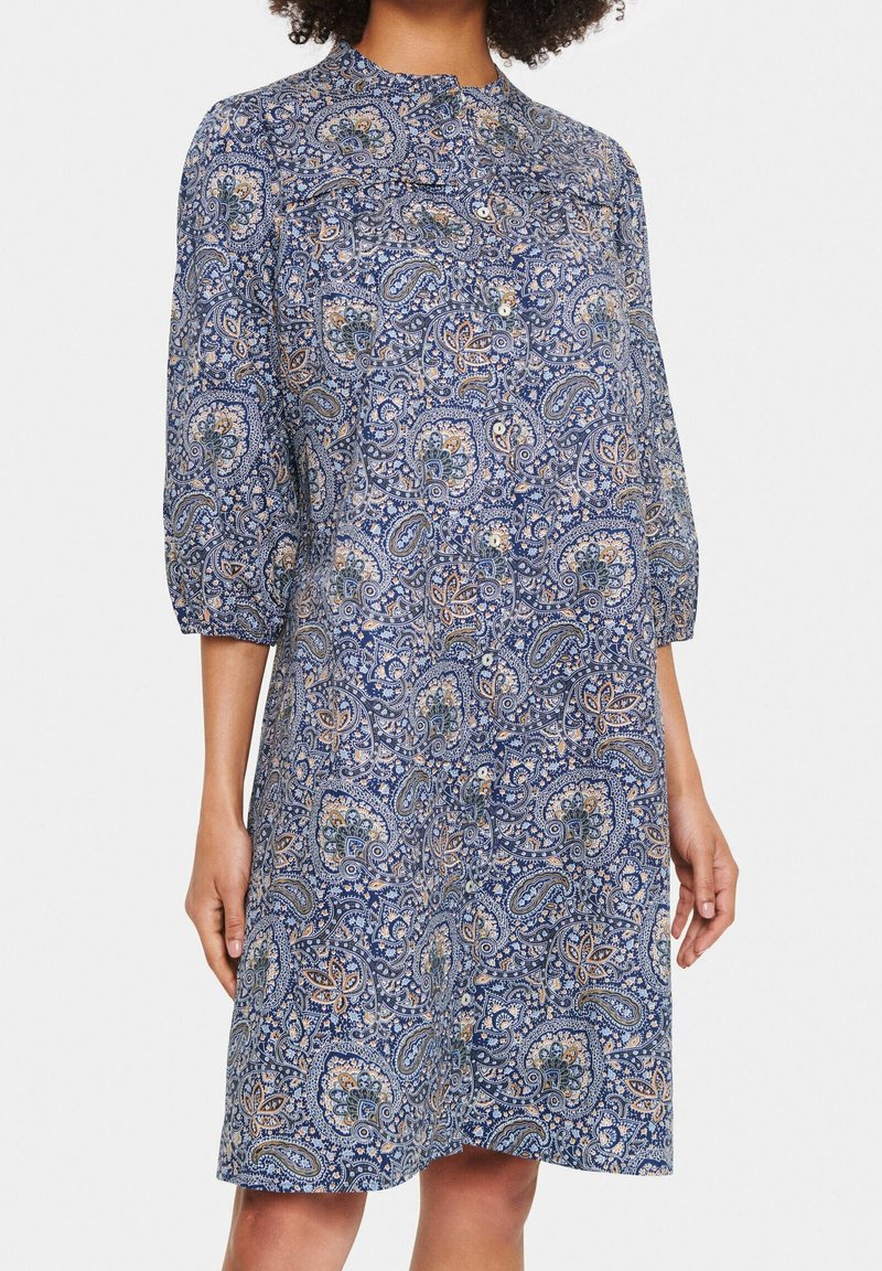 Blå paisleyklänning med 3/4 ärmar, knapprad fram och avslappnad passform. Tyget har intrikata blommönster med beige och gröna accenter.