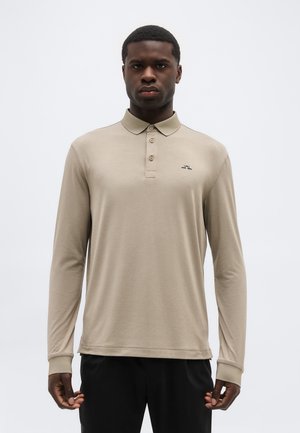 Homme portant un polo beige à manches longues avec boutons et petit logo sur la poitrine, debout devant un fond clair uni.