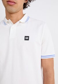 Hvid bomulds polo shirt med et sort firkantet logo patch på brystet, med blå og hvide stribede detaljer på kraven og ærmerne.