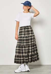 Gonna maxi a quadri nera e beige con strati sovrapposti, abbinata a una maglietta bianca e un berretto in denim. Le calzature includono sneakers bianche e calzini al polpaccio.