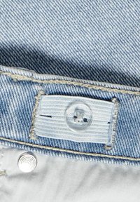 Tissu en denim bleu clair avec une fermeture à bouton visible et une étiquette blanche. Présente des coutures contrastées et un détail à bouton-pression en métal.