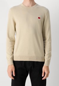 Beige Strickpullover mit rundem Hals und gerippten Bündchen. Hat ein kleines rotes Logo-Detail auf der Brust. Normale Passform, weicher Stoff.