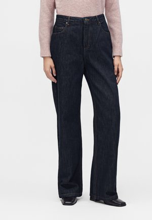 Personne portant un jean bleu foncé à jambes larges avec poches avant et un pull rose, debout devant un fond clair uni.