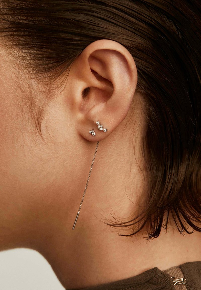 Primer plano de una oreja con tres pequeños pendientes de diamante y un pendiente de cadena de plata colgando, enmarcada por cabello corto castaño y una blusa marrón.