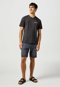 T-shirt nera in cotone con logo colorato "WRANGLER", abbinata a pantaloni corti in denim scuro e sandali neri, in piedi su uno sfondo neutro.