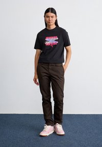 Camiseta gráfica negra con el logo colorido "PUMA Sport", pantalones marrones y zapatillas rosas con cordones blancos. Tela suave, corte relajado, diseño simple.