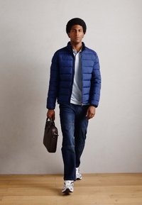Blauer gepolsterter Mantel über einem hellen Poloshirt, kombiniert mit dunklen Jeans und weißen Sneakers. Trägt eine braune Lederaktentasche. Holzboden im Hintergrund.