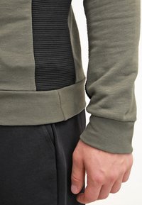 Olijfgroene langemouw shirt met een gestructureerd zwart paneel, geribbelde accenten aan de zijkant en een strakke manchette, gecombineerd met donkere broek.