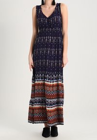 Mouwloze marineblauwe maxi-jurk met V-hals, bedekt met beige en meerkleurige geometrische patronen, gedragen met zwarte instap schoenen.