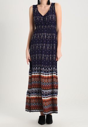 Ärmelloses maximales Kleid in Marineblau mit V-Ausschnitt, gemustert mit beigen und bunten geometrischen Designs, getragen mit schwarzen Slipschuhen.