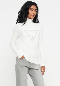 Donna con lunghi capelli neri che indossa un maglione a collo alto bianco con un logo "GUESS" tempestato di gioielli e jeans grigio chiaro, in piedi contro uno sfondo bianco.
