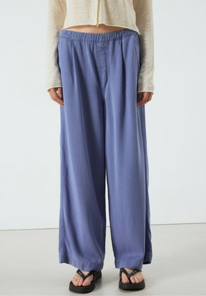 Person trägt lockere blaue Hose, schwarze Plateau-Zehentrenner und ein kurzes hellbeiges Langarmoberteil, steht drinnen auf einem schlichten Boden.