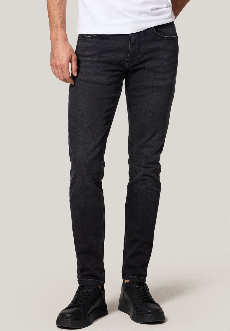 Mężczyzna w dopasowanych czarnych dżinsach, zestawionych z czarnymi sneakersami i białą koszulą, stojący na tle jednolitego, jasnego tła.