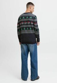 Pleteni pulover z črnim podlagom, ki ima v ponavljajočem se vzorcu zelene drevesa in rdeče okraske, v kombinaciji z ohlapnimi modrimi jeans hlačami.