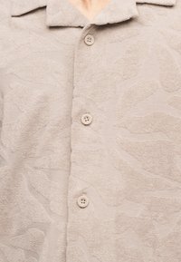 Μπεζ υφασμάτινο πουκάμισο με διακριτικό αφηρημένο σχέδιο, κουμπωμένο μπροστά και με φαρδύ γιακά ορατό στο λαιμό.