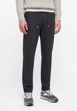 Homme portant un pantalon de jogging gris foncé avec cordons, des baskets grises et blanches, et un pull en maille gris clair, visible de la taille aux pieds.