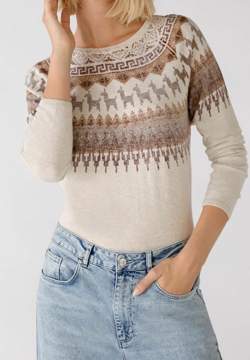 Pull beige avec des motifs géométriques marron et gris et des motifs d'animaux, col rond et manches longues ajustées, associé à un jean bleu clair.