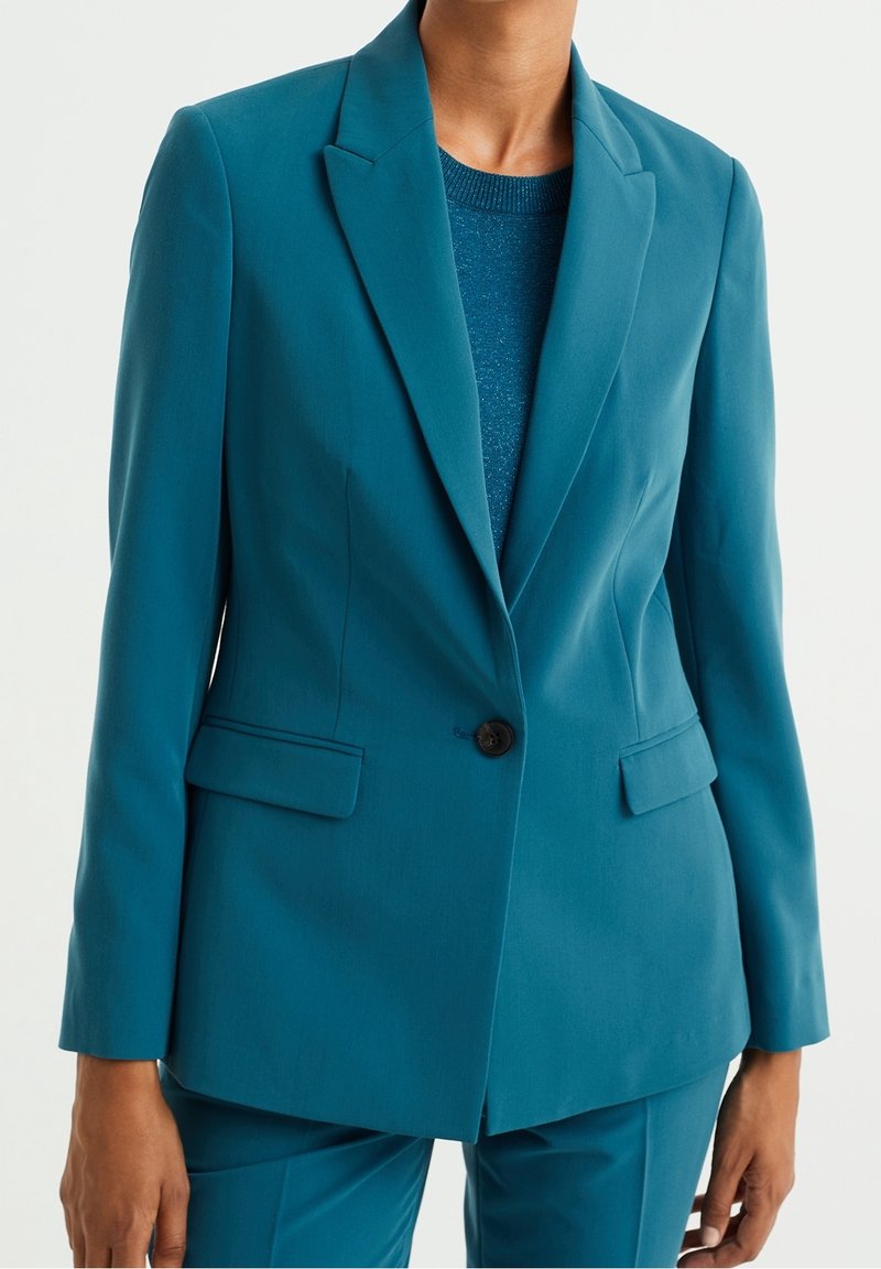 Blazer sur mesure bleu sarcelle avec un bouton noir, poches à rabat, porté sur un haut en maille bleu sarcelle assorti et un pantalon bleu sarcelle, sur une personne devant un fond clair.