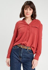 Chemise boutonnée de couleur rouille avec deux poches avant, tissu texturé et col, assortie à un jean bleu taille haute et une ceinture à imprimé léopard.