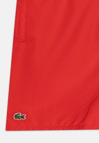 Articol vestimentar roșu, confecționat dintr-un material fin, având un mic logo brodat cu un crocodil verde în partea de jos. Designul este minimalist, fără modele.