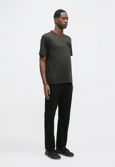 Tricou din bumbac negru cu un design floral texturat pe partea stângă a pieptului, asortați cu pantaloni negri și pantofi negri cu bretele.