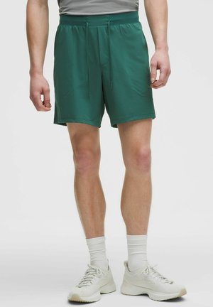 Zöld zsinóros sportnadrágot, fehér zoknit és fehér sportcipőt viselő férfi, egy egyszerű világos háttér előtt áll.