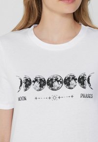 Camiseta blanca de algodón con un estampado gráfico negro de las fases de la luna y estrellas, con el texto "MOON" y "PHASES" debajo del diseño.