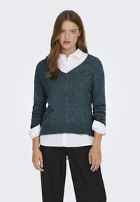 Dunkeltürkisfarbener V-Ausschnitt-Pullover aus weichem Strick mit langen Ärmeln und geripptem Saum, über einem weißen Hemd mit Kragen getragen und kombiniert mit schwarzen Hosen.