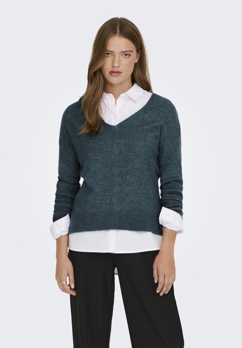 Dunkeltürkisfarbener V-Ausschnitt-Pullover aus weichem Strick mit langen Ärmeln und geripptem Saum, über einem weißen Hemd mit Kragen getragen und kombiniert mit schwarzen Hosen.