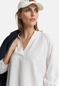Witte linnen blouse met een V-hals en een relaxte pasvorm. Beschikt over pofmouwen en een subtiele textuur. Vergezeld van een marineblauwe jas en een crèmekleurige pet.