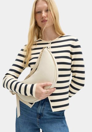Kvinde med langt blondt hår iført en sort og hvid stribet sweater og blå jeans, der holder en beige håndtaske i halvmåneform.