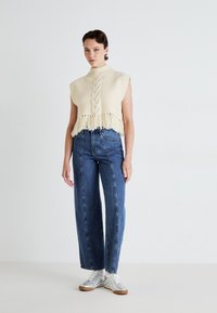 Kremfarget, kort genser med mock neck, flette- og frynse detaljer; sammen med høyt liv blå denimbukser og sølvfargede sneakers.