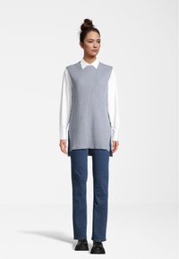 Un pull sans manches en tricot côtelé gris clair sur une chemise blanche à col, associé à un jean droit bleu foncé et des chaussures noires.