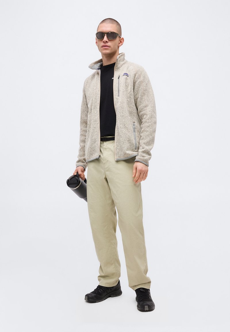 Man wearing beige fleece jacket, black sunglasses, black shirt, beige pants, and black shoes, holding a black reusable water bottle.