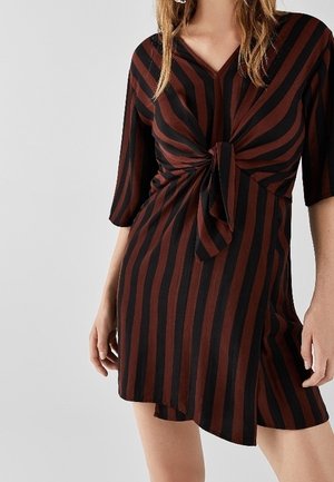 Frau trägt ein kurzes braun-schwarzes Kleid mit vertikalen Streifen, einem Knotendetail vorne und ellbogenlangen Ärmeln.
