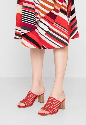 Jambes portant des sandales rouges à talons hauts ouverts avec des talons épais en bois et une jupe colorée rayée arrivant au genou.